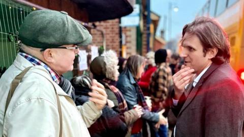 North Norfolk MP Steff Aquarone talking to protesters and residents at the Sheringham Bus Shelter. He is on the right and is wearing a suit and is deep in conversation with a man, standing on the left, who is wearing a coat, a chequered shirt and a flat cap.
