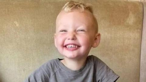 Three-year-old Albie Speakman, with light blond hair and a beaming smile, is photographed in a grey t-shirt.