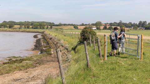 Two walkers walk through a gate that connects two fields. There is a river on the left hand side. In the distance there are some buildings and further fields. 