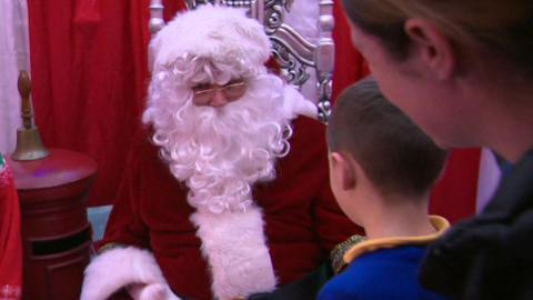 Father Christmas, wearing a red and white coat, is sat on a silver chair talking to a boy with brown hair and a woman with dark hair.