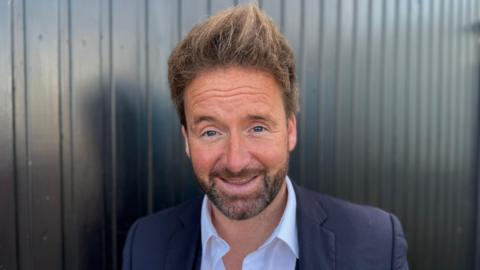 A man with light brown hair and a dark beard wearing a white shirt and a suit jacket.