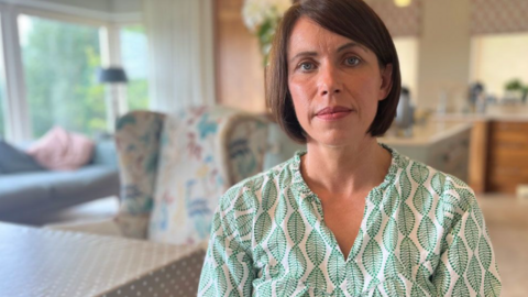 Angela O'Neill, a woman with brown, bobbed hair, looks into the camera.  She is wearing a white top patterned with green leaves.  There is a blurred background of a kitchen-diner behind her, with a large window on the left overlooking trees. 