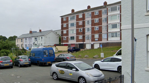 A street view image of Stonehall Flats. The block of apartments is brown with white stripes. Outside is a filled car park.