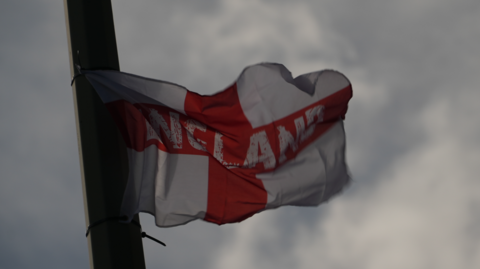 A St George's Cross flag - a white rectangular piece of fabric with a red cross on it from top to bottom and left to right. England is printed across the horizontal stripe in white lettering. It is fluttering in the breeze, having been attached to a lamppost by cable ties. The sky behind it is cloudy and grey.