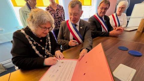 Truro city mayor Carol Swain, wearing a black top and her mayoral chains, signs a twinning partnership on a table with Morlaix mayor Jean-Paul Vermot, who is wearing a grey suit and a sash containing the colours of the French flag - red, white and blue.