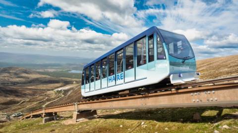 One of the funicular's cars heads uphill along rails laid on the mountainside. It is called Hare and its name is on the side of the vehicle, which is different shapes of blue. There are clouds in the sky.