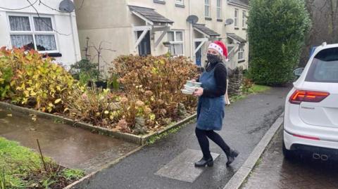 Sue Clarke outside of a house wearing a red Christmas hat with a blue pinafore and black. She is walking towards a house with Christmas meals in her hands. The garden has bushes and flowers in the front. 