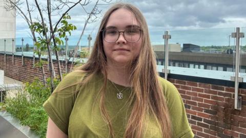A young woman stands in front of a low-brick wall and a planter. She has long brown hair and wears glasses, a green t-shirt and a silver necklace with a large pendant on it.