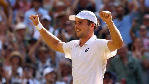 Arthur Ferrt celebrating his win with his arms in teh air and smiling at Wimbledon with spectators blurred in the background