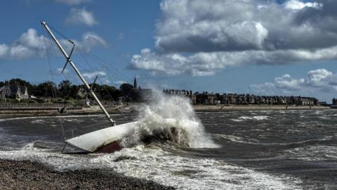A large wave crashes against a yacht which tips to the side with a cloudy sky and buildings on a sea shore in teh background