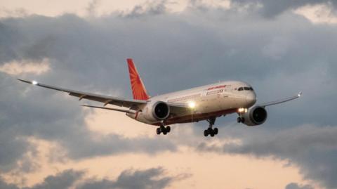 Stock picture of a large twin-engine aircraft in flight against a sky with light clouds. Its livery is the red and white of Air India 