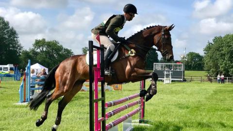 A horse and rider jump over a pink, black and white jump in a field with other jumps in. The horse is brown with black main and tail, the rider wears a green jacket and beige jodhpurs with a black helmet