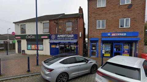 The store can been next to other businesses on the road. It is a lower ground business with string lighting hanging down from the floor above. The shop signage is blue.