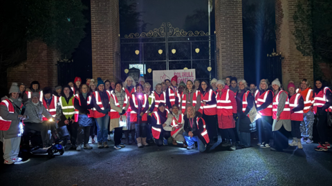A large group of women together outside a park at night ahead of a walk. they are all wearing orange hi vis jackets