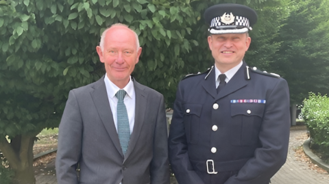 Simon, on the right, wears a police uniform and a police cap standing next to Darryl, who is wearing a grey suit, white shirt and a green tie.