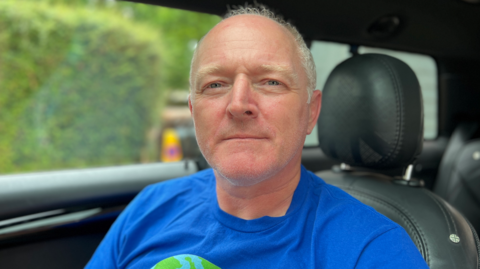 Mike Nunn is sitting in the driving seat of his car. He is wearing a blue t-shirt. Mike has grey hair and blue eyes. In the window of the car a blurry view of a garden bush can be seen.