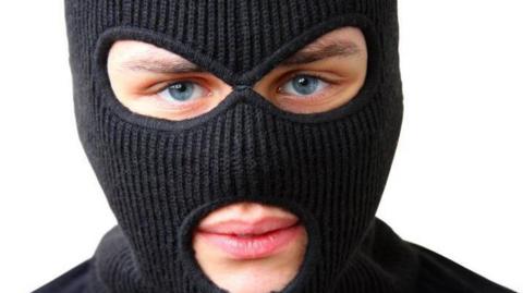 A close-up of a man wearing a black, full-face balaclava. He is standing in front of a white background.