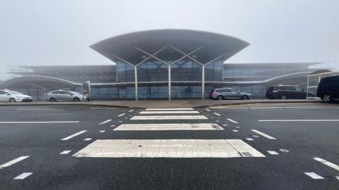 A zebra crossing in front of a small but modern airport terminal building on an overcast day.