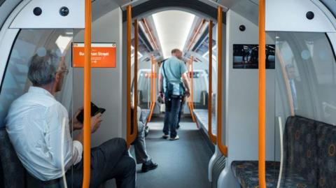 The inside of a Glasgow Subway. A man sits on one of the seats which face each other along the long corridor of the vehicle. He looks at his phone and sits next to a sign which says the next stop is Buchanan Street