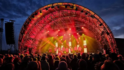 A dome-like structure stands over the stage with red and yellow lights. The night sky can be seen behind it with a crowd of people stood in front of the stage.