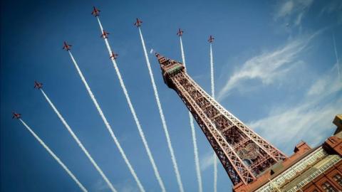 The Red Arrows fly over Blackpool Tower leaving trails of white smoke against a blue sky. 