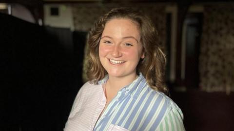 Alexandra Godfrey smiling at the camera. She is a young woman with a blue and pink striped shirt on and has curly brown hair.  The background is dark because she is at a silent disco. 