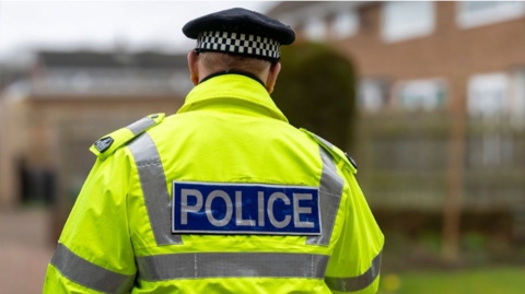 The back of a police officer in a high-visibility neon yellow jacket with the word "Police" on the back.