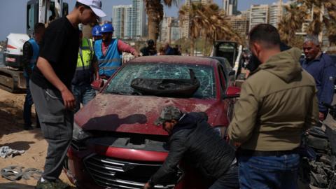 Lebanese officials and civilians inspect a car damaged in an Israeli strike on Ramlet al-Baida beach in Beirut, Lebanon (12 March 2026)