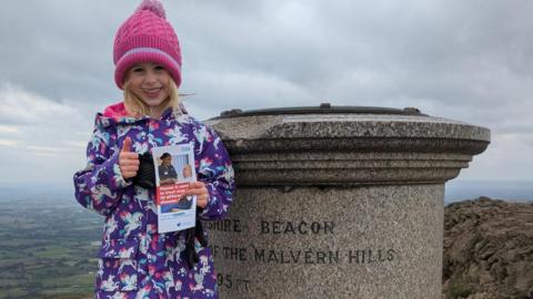 A young girl with blonde shoulder-length hair smiles as she stands at the top of the Worcestershire Beacon, next to the marker at the top, holding an NHS leaflet with information on about plasma donation. She is giving a thumbs up with her other hand and is wearing a pink beanie hat and a purple coat with unicorns on.