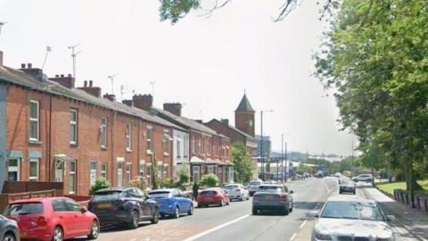 Parked cars and red-bricked terraced houses are on the left hand side of the road with a grassed area and trees on the right hand side.