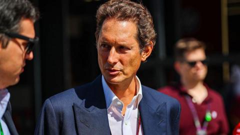 A man with brown hair wearing a navy blue suit jacket and white shirt stands in bright sunlight, talking to another man wearing sunglasses off to his right.