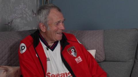 Tim Goodchild with short grey hair seated indoors on a sofa. He is wearing a red, black, and white sports jersey. The background is softly blurred, showing grey-toned furnishings and a wall, with him facing toward the right side of the image.
