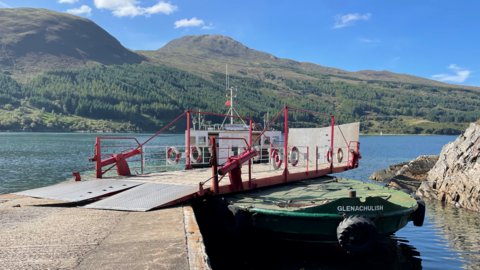 Bàta-aiseig beag dearg agus uaine le clàr-tionndaidh leis an ainm Glenachulish sgrìobhte oirre. Tha i ceangailte ri lamraig. Tha am muir ciùin agus tha a' ghrian a' deàrsadh. Tha cnuic le craobhan-giuthais air a cùlaibh. 