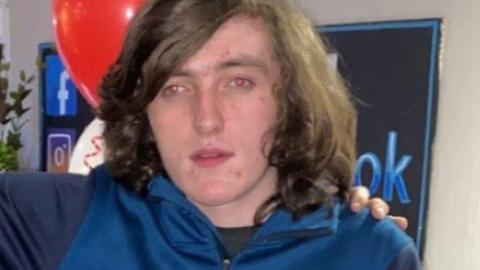 Nineteen-year-old Nyle Corrigan, with shoulder-length brown hair and wearing a blue zip-up top, poses for the camera in front of a red balloon.