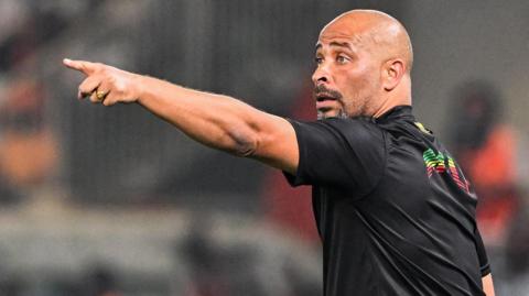 Eric Chelle, wearing a black T-shirt and with his back half turned to camera, points his left arm out in front of him as he gestures during a football match