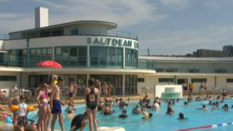 Saltdena Lido. There is an outdoor swimming pool with lots of swimmers in. In the background is a white paviliuon-style building with the words Saltdean Lido writen in blue on it. The sky is blue with clouds