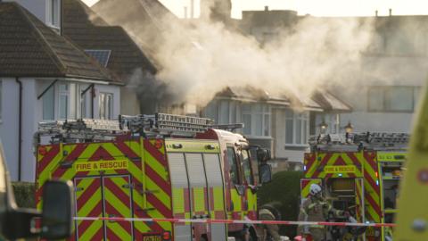 Fire engines and firefighters outside a home that has smoke coming from an upstairs window.