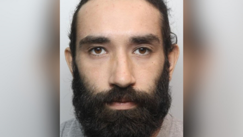 Police mugshot of Usama Al-Sari, who is a 28-year-old man with a full dark beard. Wearing a grey t-shirt, he stares impassively towards the camera.