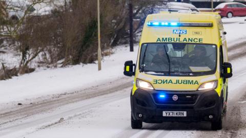 A yellow ambulance with its blue emergency lights on is driving along a snowy road. The grey tarmac is only peeking through where other cars have driven along it.