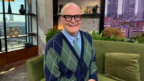 Lawyer Gary Rycroft is sitting on a green velvet sofa in the Morning Live studio, wearing a green and blue cardigan, with a green tie and blue shirt underneath. He is wearing black framed glasses and is smiling at the camera