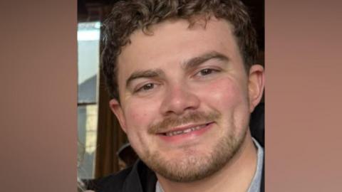 A bearded man wearing a grey t-shirt and jacket.