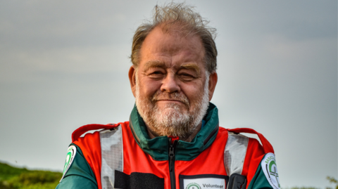 Dr David Hillebrandt, looking directly at the camera, is wearing a red hi-vis jacket and green jacket. He has a name badge which says "Volunteer" on it. The sky behind him is grey.