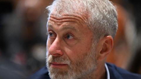 A close up of Roman Abramovich looking away from the camera to the left. He is wearing a blue suit and white shirt and has a grey and white beard.
