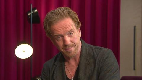 Damian Lewis looks directly at the camera with his eyebrows raised. He is wearing a dark linen jacket and behind him is a red curtain and black lamp.