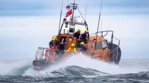 A wide shot of the RNLI all-weather lifeboat. The waves are crashing around the orange and black vessel, and there are multiple members of crew on board.