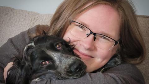 Emily Thornton-Sandy sitting on a sofa with a dog. She is hugging it to her chest and the dog had his snout on her arm. It has black fur with mottled white on the top of his nose going up between his eye. She has a broad smile and light brown hair down to her shoulders. She is wearing a grey top and glassed with a black rim on top 