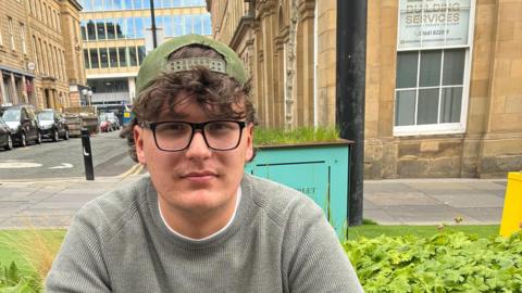 Cameron Scott. He has brown hair and is wearing a green cap and a jumper. He is sitting on Grey Street in Newcastle with an area of green behind him an large brown office buildings of various ages behind him.