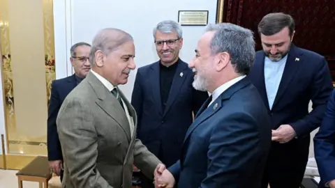Pakistan's Prime Minister Shehbaz Sharif (L) shakes both hands of Iranian Foreign Minister Abbas Araghchi (R). Both are wearing suits, three other officials observe them from the side 