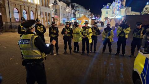 Several police officers, standing in a semi-circle in Cathedral Square in Peterborough in the evening, with one officer standing before them and addressing them.