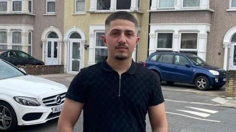 A man in a black shirt with a zip on the front stands in a street. He has short dark hair and a beard.